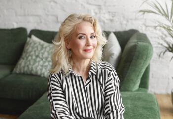 Beautiful middle aged 50 year old female wearing stylish elegant striped blouse relaxing at home, sitting on comfortable green sofa in modern living room interior, looking at camera with joyful smile