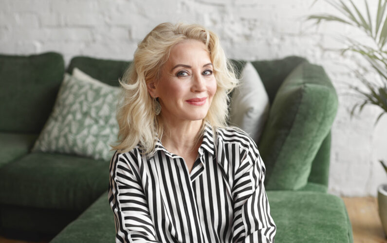 Beautiful middle aged 50 year old female wearing stylish elegant striped blouse relaxing at home, sitting on comfortable green sofa in modern living room interior, looking at camera with joyful smile